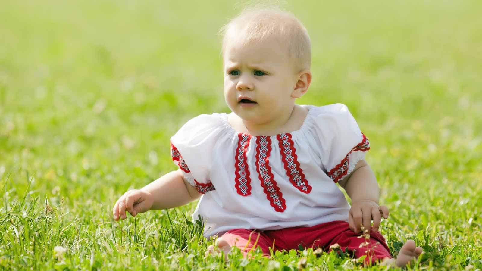 A baby with light hair wearing a white shirt with red embroidery and red pants sits on green grass outdoors.