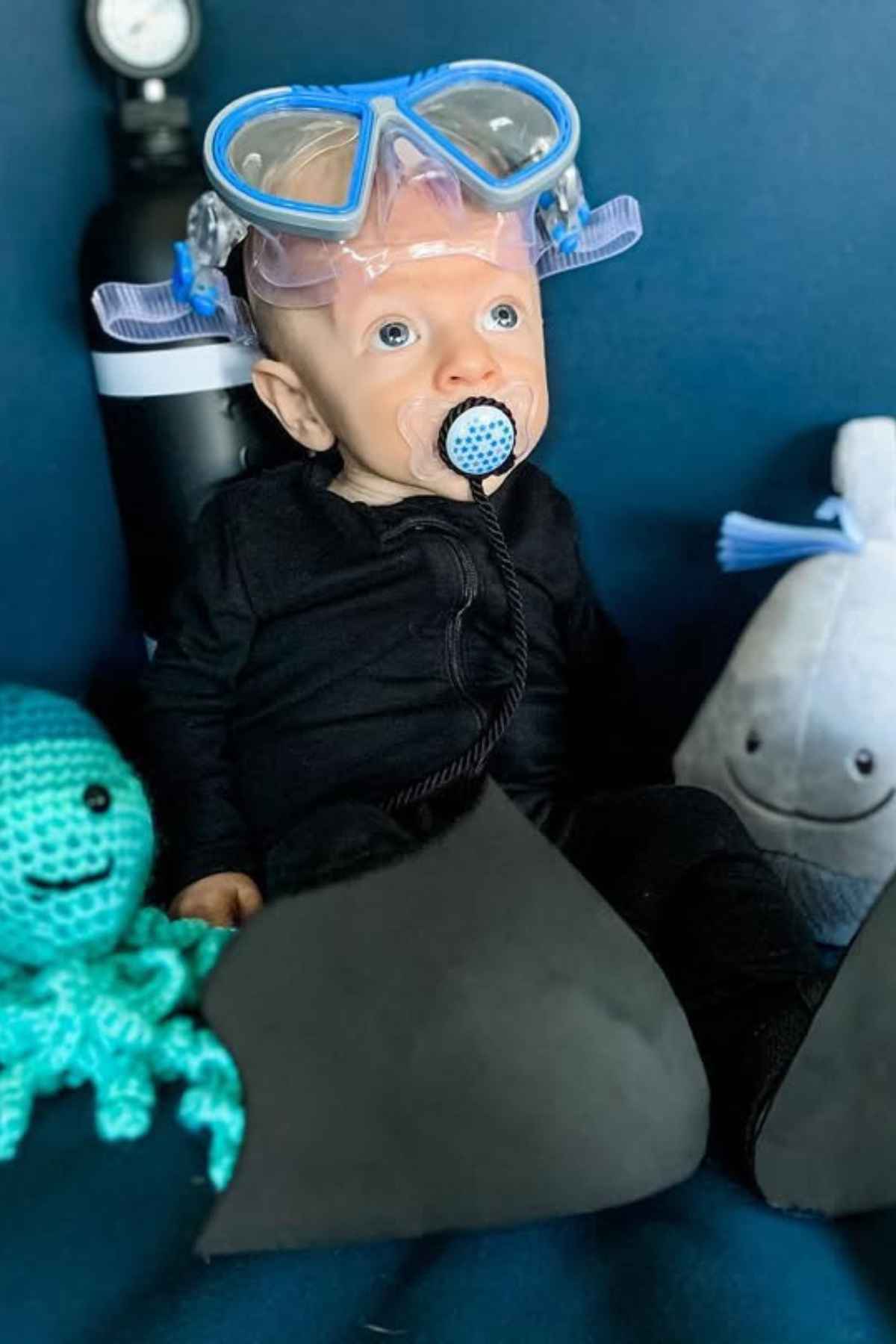Baby dressed as a scuba diver, with a pacifier on mouth, goggles places on forehead, oxygen tank at the bank and an octopus and shark toys beside him.