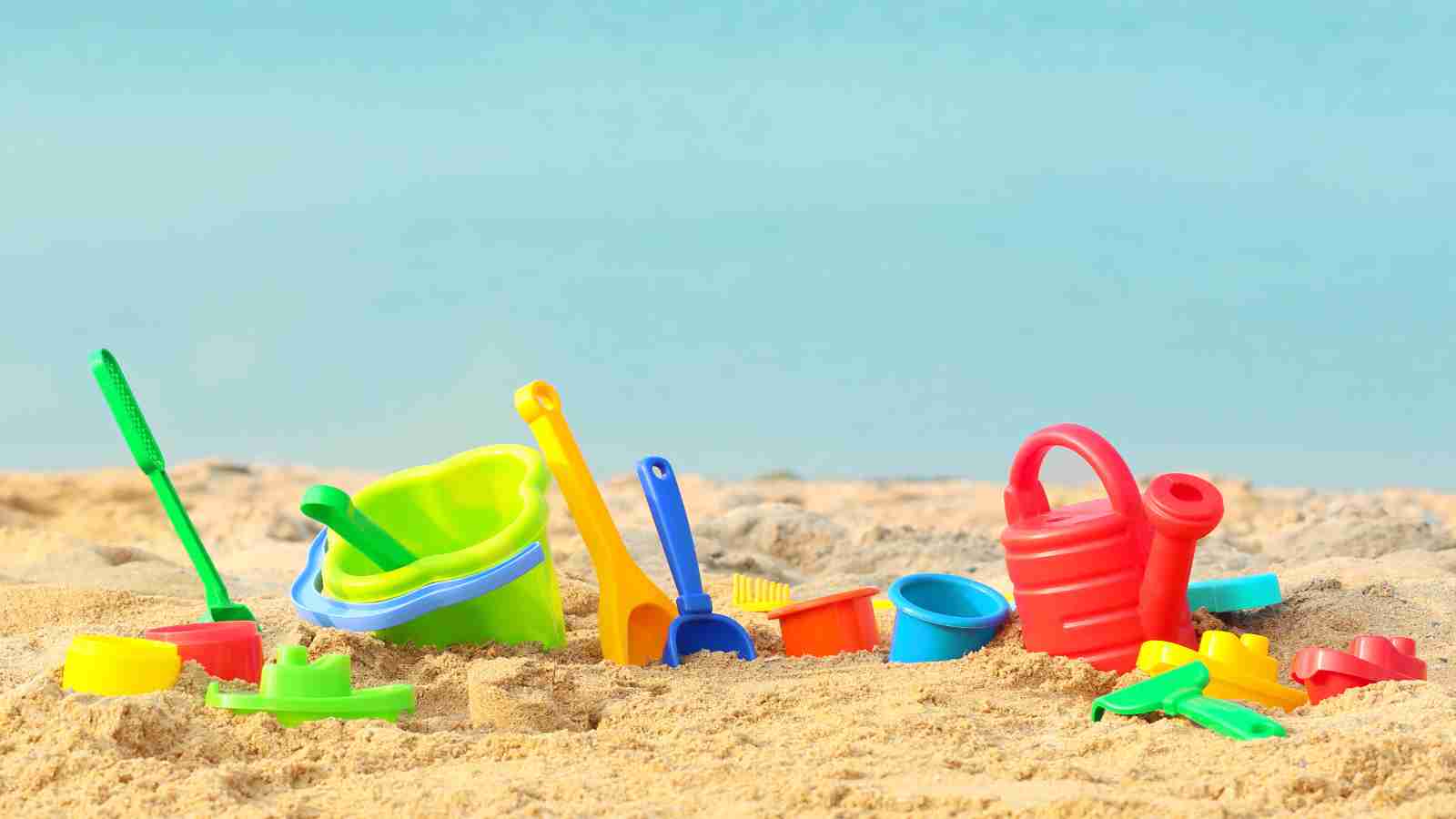 Colorful plastic beach toys, including buckets, shovels, and a watering can, scattered on sandy beach with calm blue water in the background.