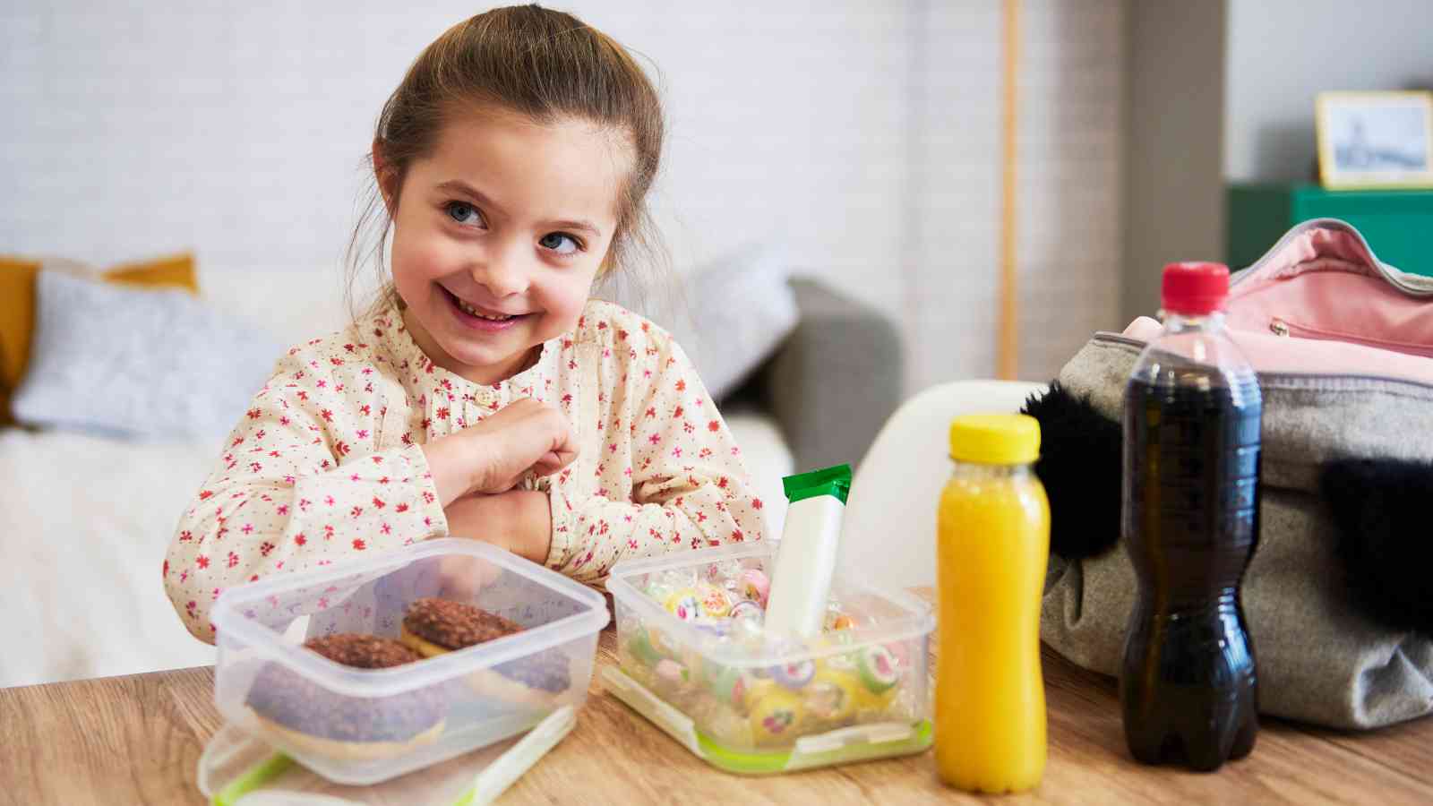A smiling young girl sits at a table with her lunchbox, containing cookies and candy, and drinks, ready for school.