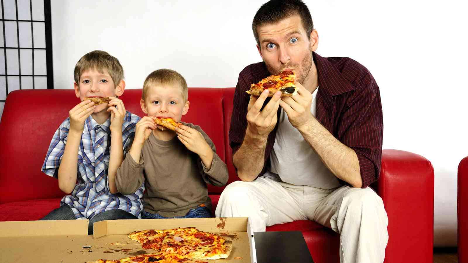 A father and his two sons happily eat pizza together while sitting on a red couch.