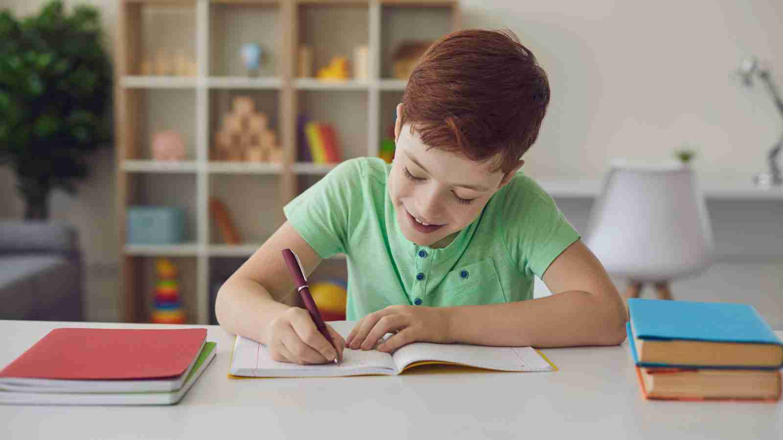A smiling boy with red hair sits at a table, writing in a notebook with a pen.