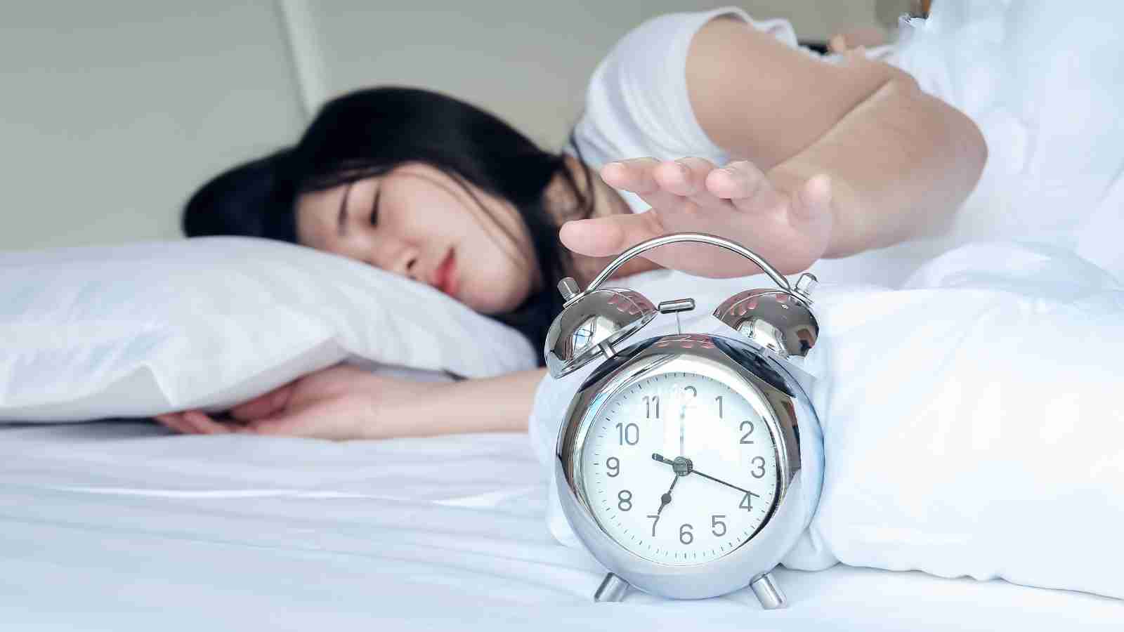 A woman sleeps in bed, her hand reaching out to turn off a ringing alarm clock.