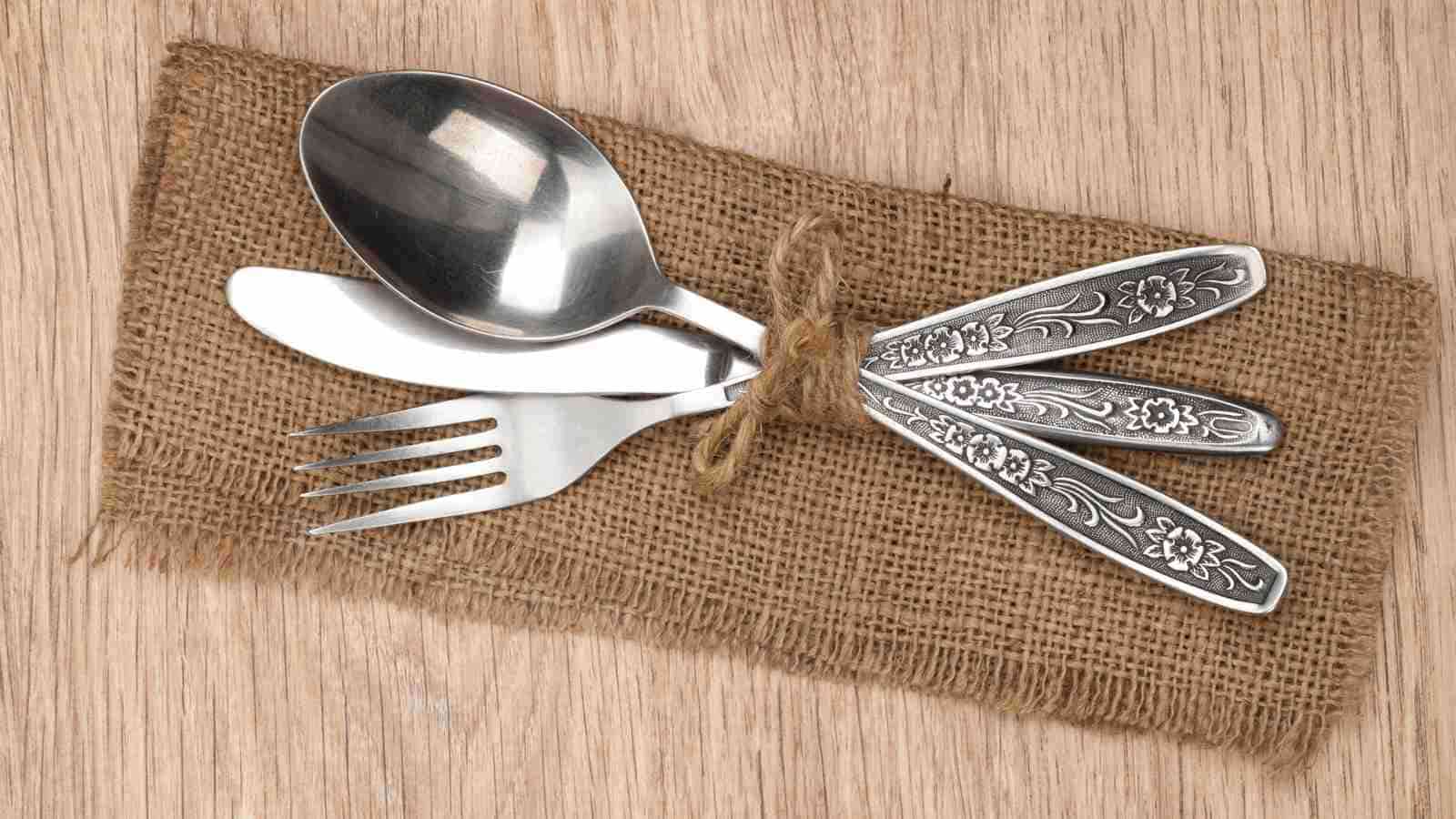 A spoon, fork, and knife with ornate floral designs are tied together with twine and placed on a burlap napkin.