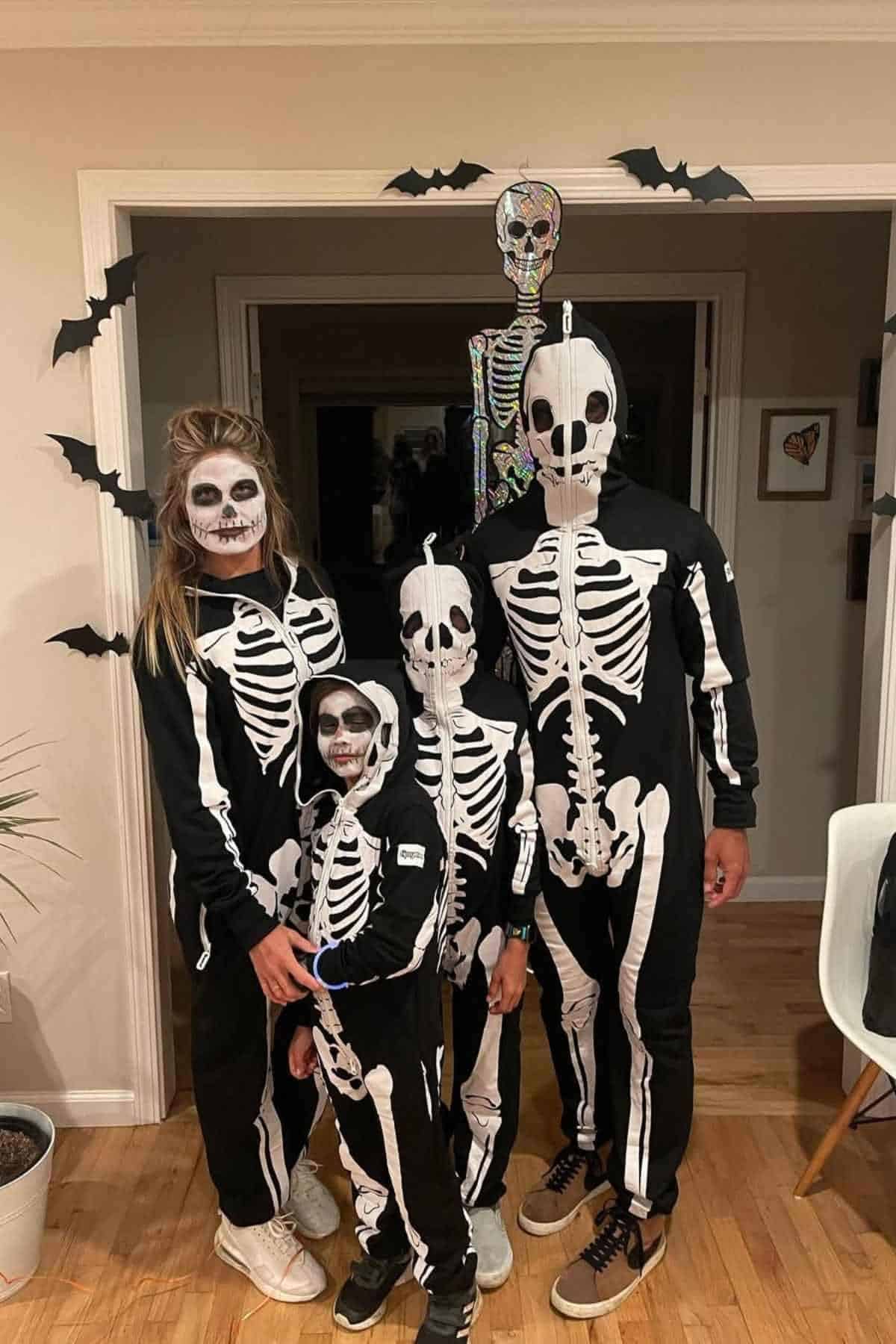 Four people wearing matching black and white skeleton costumes stand together indoors, with Halloween decorations including bats and a skeleton figure in the background.