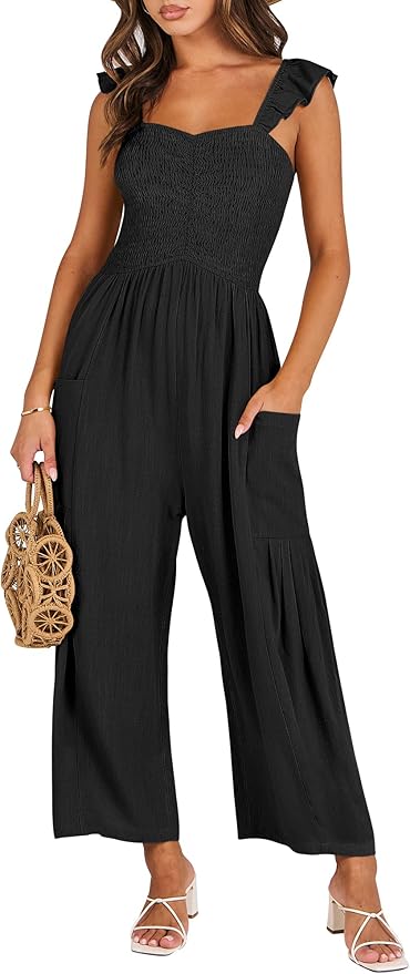 Woman wearing a sleeveless black jumpsuit with ruffled straps, wide legs, and pockets, holding a woven handbag and wearing white heeled sandals.