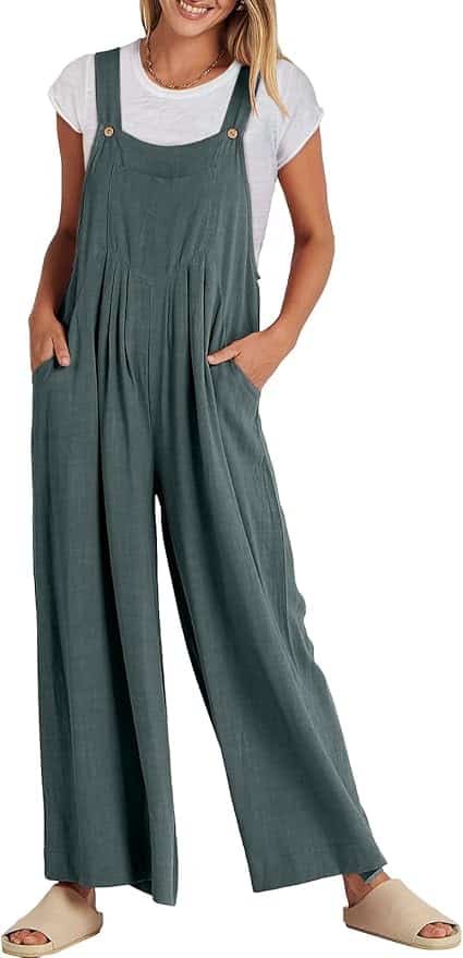 Woman wearing a white t-shirt under loose-fitting, sleeveless green overalls with wide legs and beige slip-on shoes. Hands are in the overalls pockets.