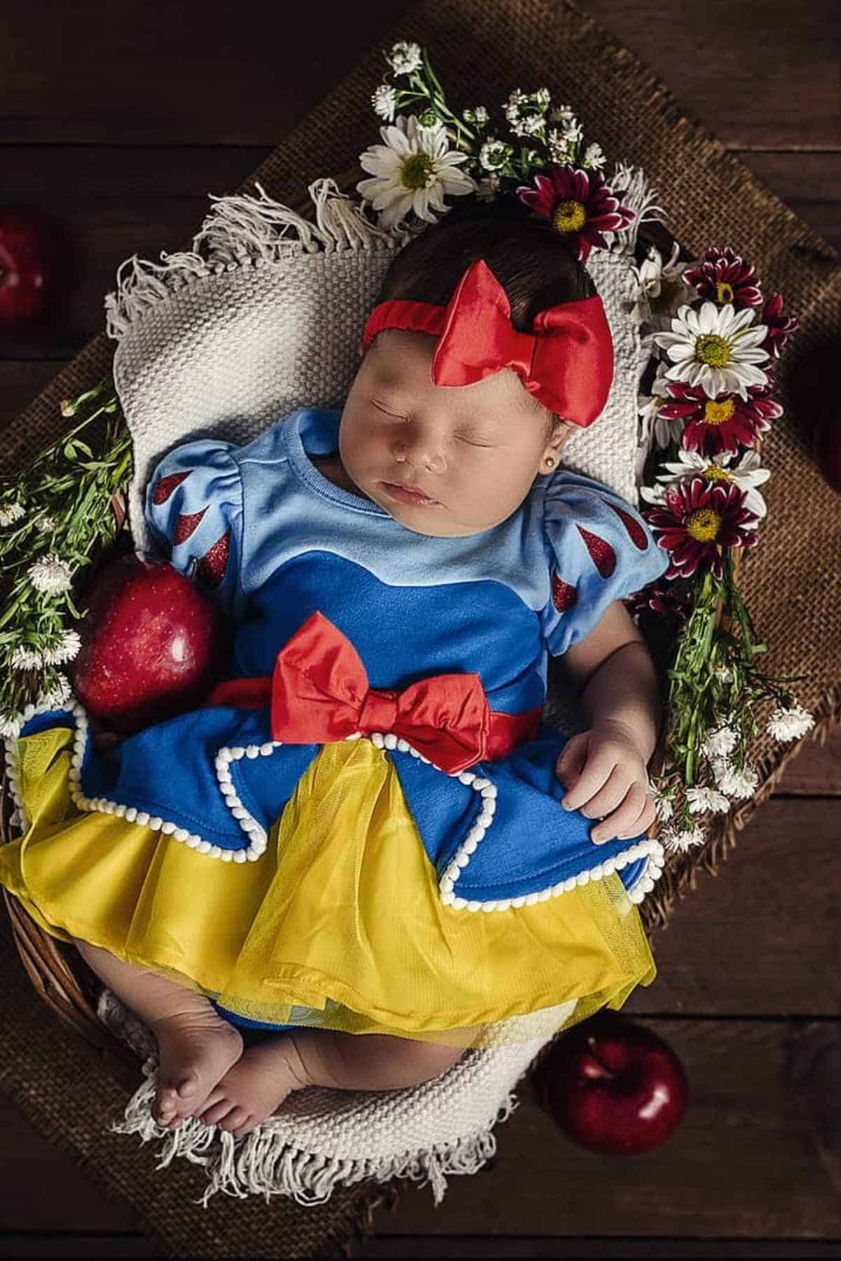 A baby dressed in a Snow White costume with a red bow headband sleeps in a basket decorated with flowers and surrounded by red apples on a wooden surface.
