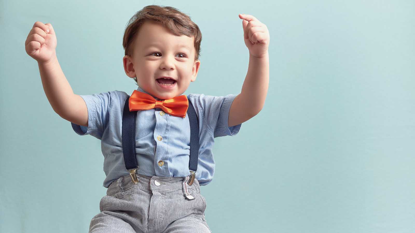 A baby boy wearing a blue short sleeve, orange bow tie and patterned pants with suspenders.