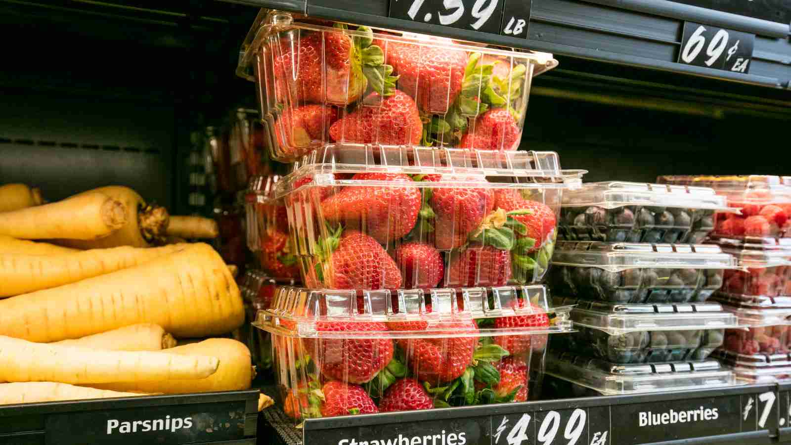 A grocery store shelf displays containers of strawberries, blueberries, raspberries, and parsnips with price tags.