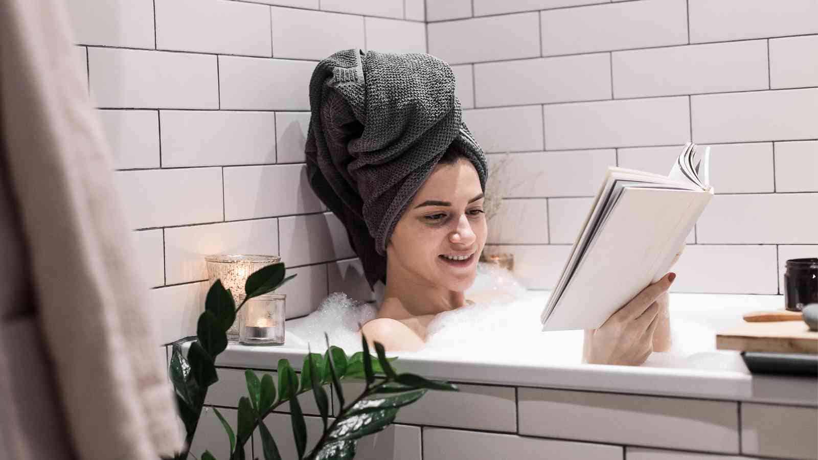 A woman relaxes in a bubble bath, reading a book while surrounded by candles and plants.