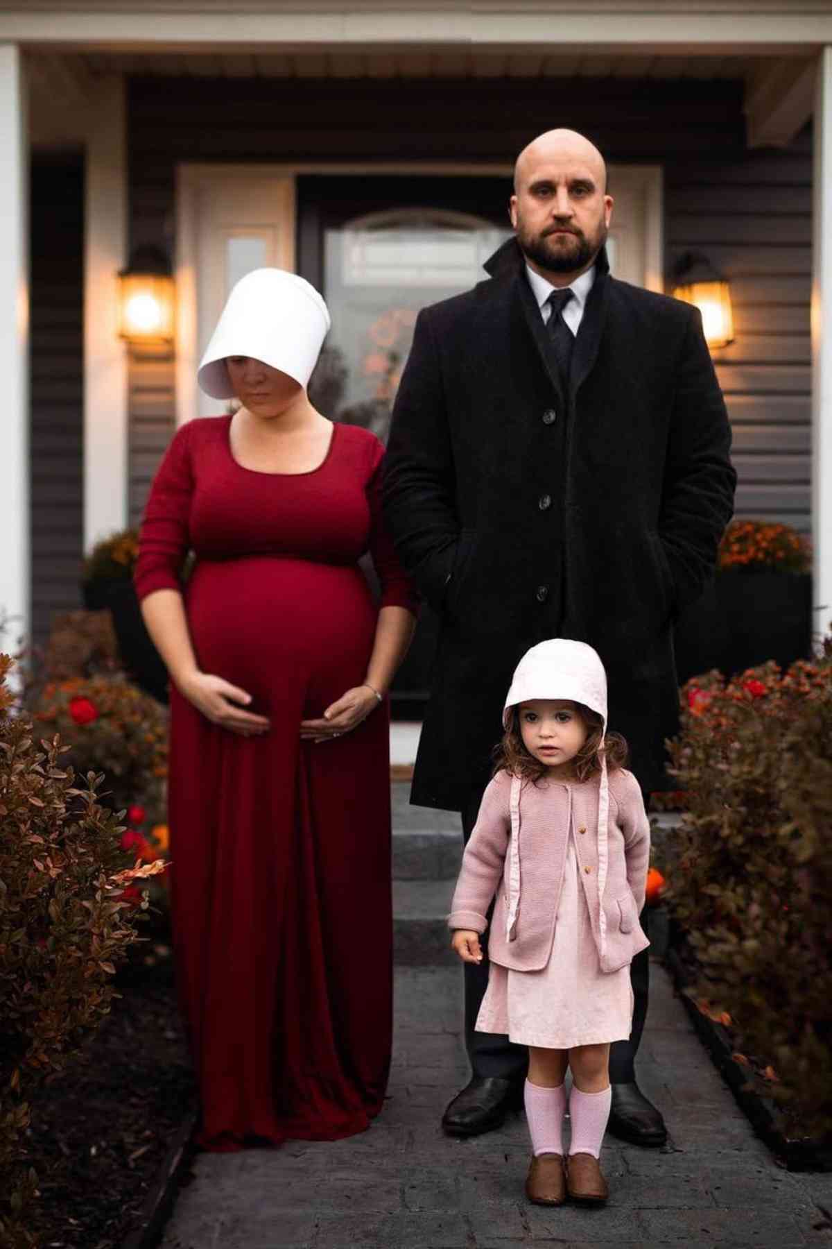 A pregnant woman in a red dress, a man in a black coat and tie, and a young girl in a pink coat stand outside a house, facing the camera.
