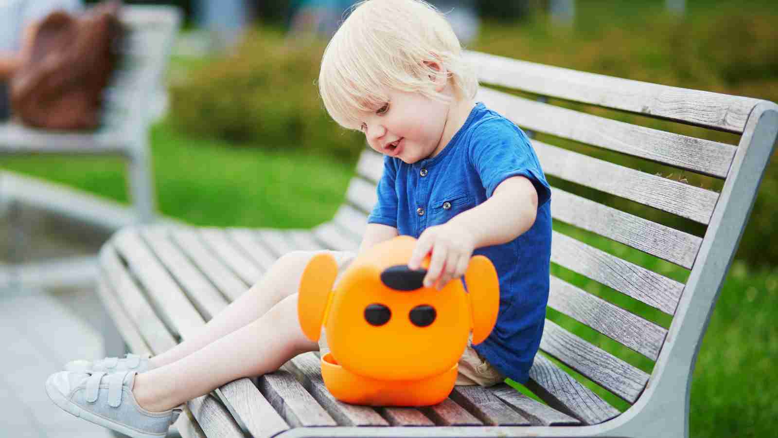 Young child wearing a blue shirt sits on a wooden bench outdoors, interacting with an orange robot toy.
