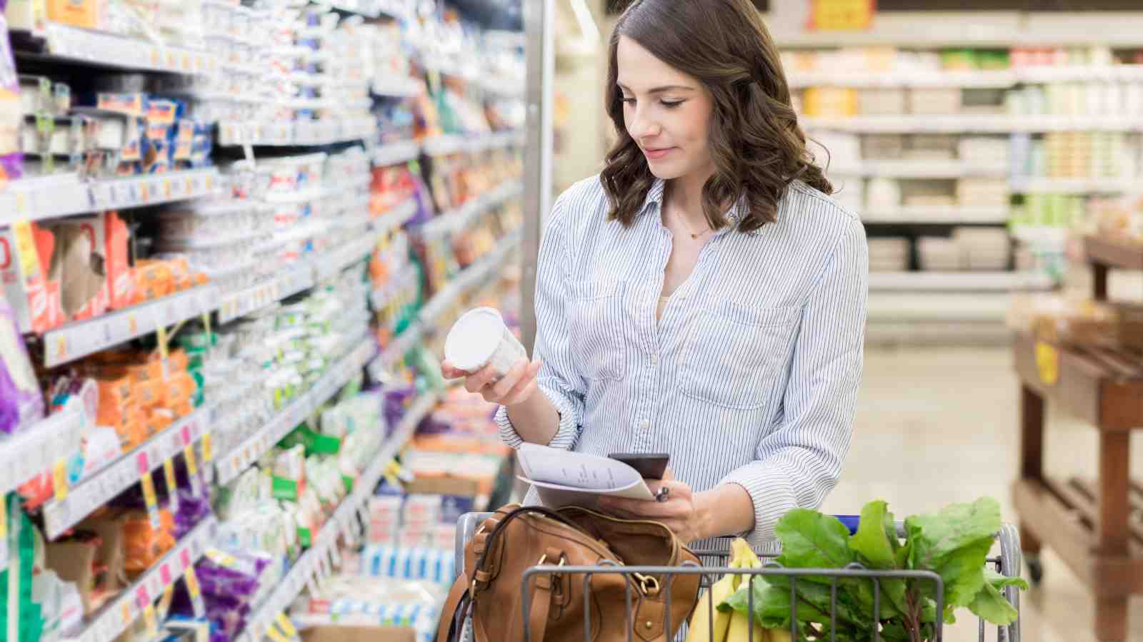 A woman examines a yogurt container in a grocery store while holding a shopping list and pushing a shopping cart.