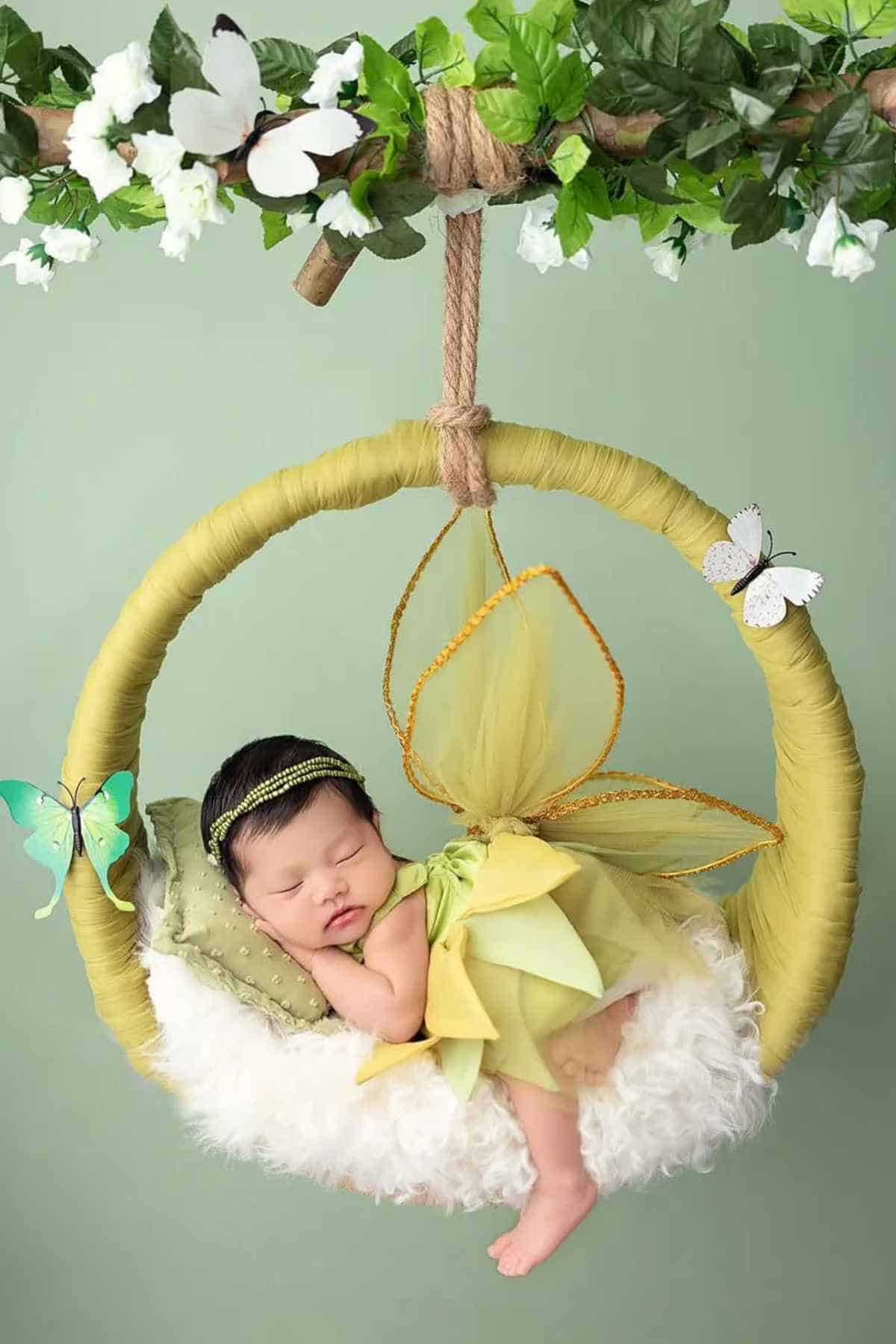 A baby dressed in a green fairy costume sleeps on a fluffy cushion inside a circular swing decorated with butterflies and greenery.