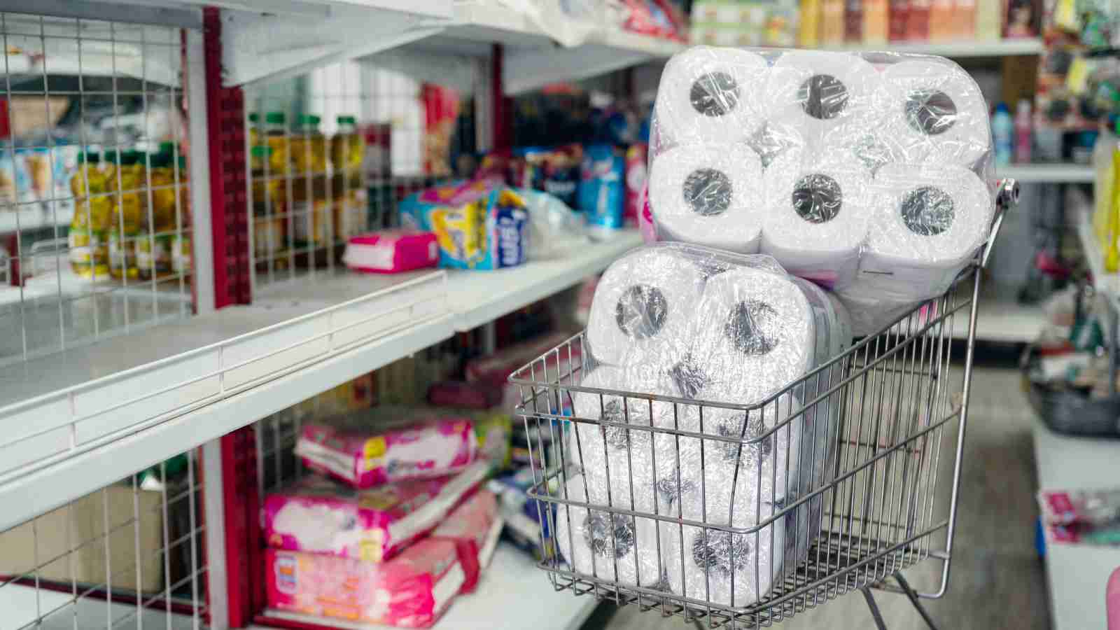 A shopping cart filled with packages of toilet paper stands in an aisle with sparsely stocked shelves.
