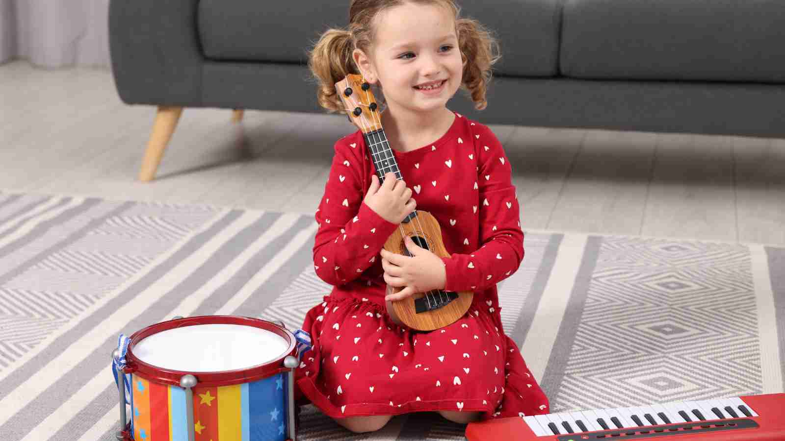 Young girl in a red dress with white hearts sits on a rug holding a small guitar, with a toy drum and toy keyboard in front of her.
