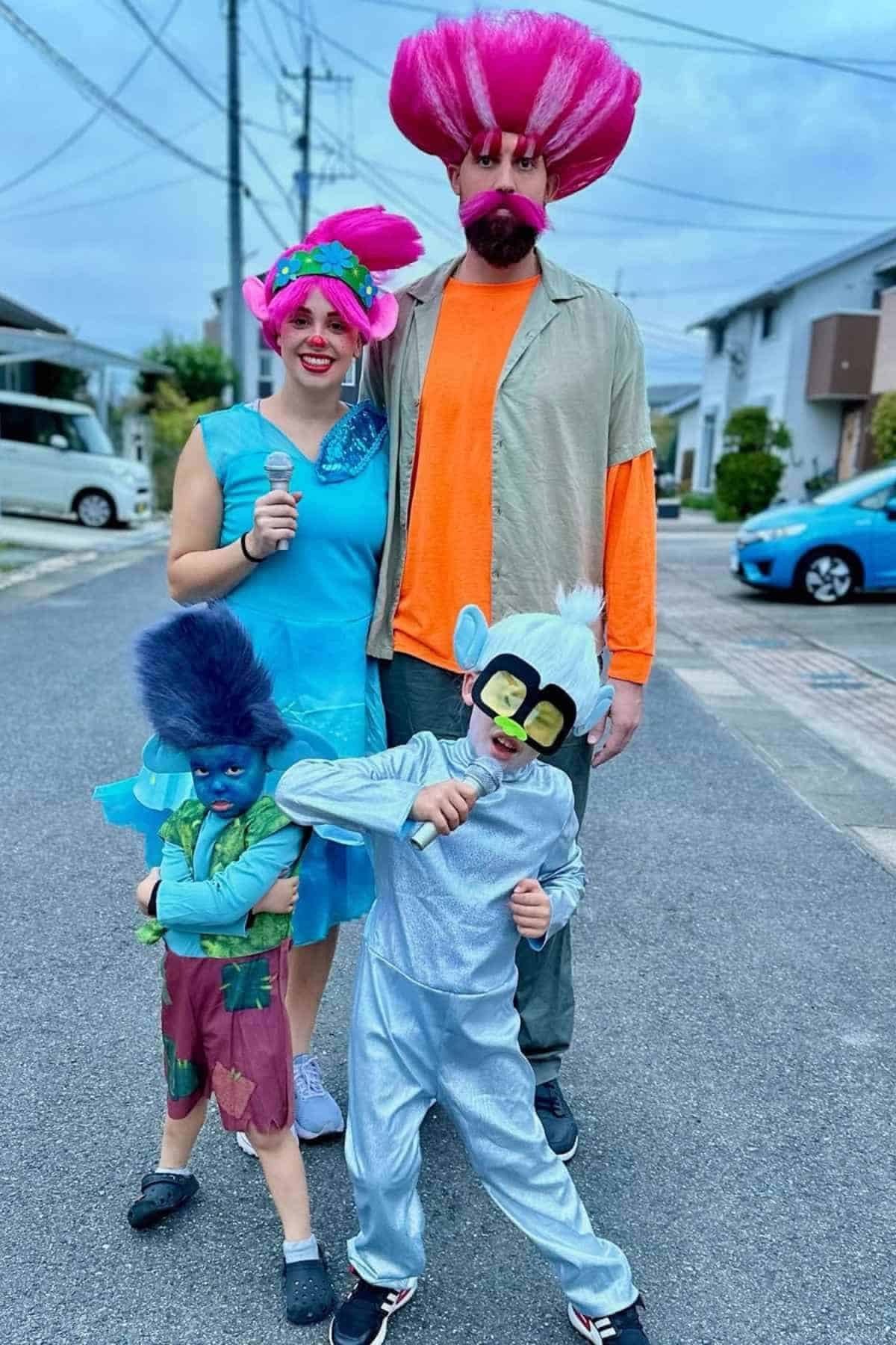 Four people wearing colorful Trolls costumes stand together on a residential street, dressed as characters with vibrant wigs and themed outfits.