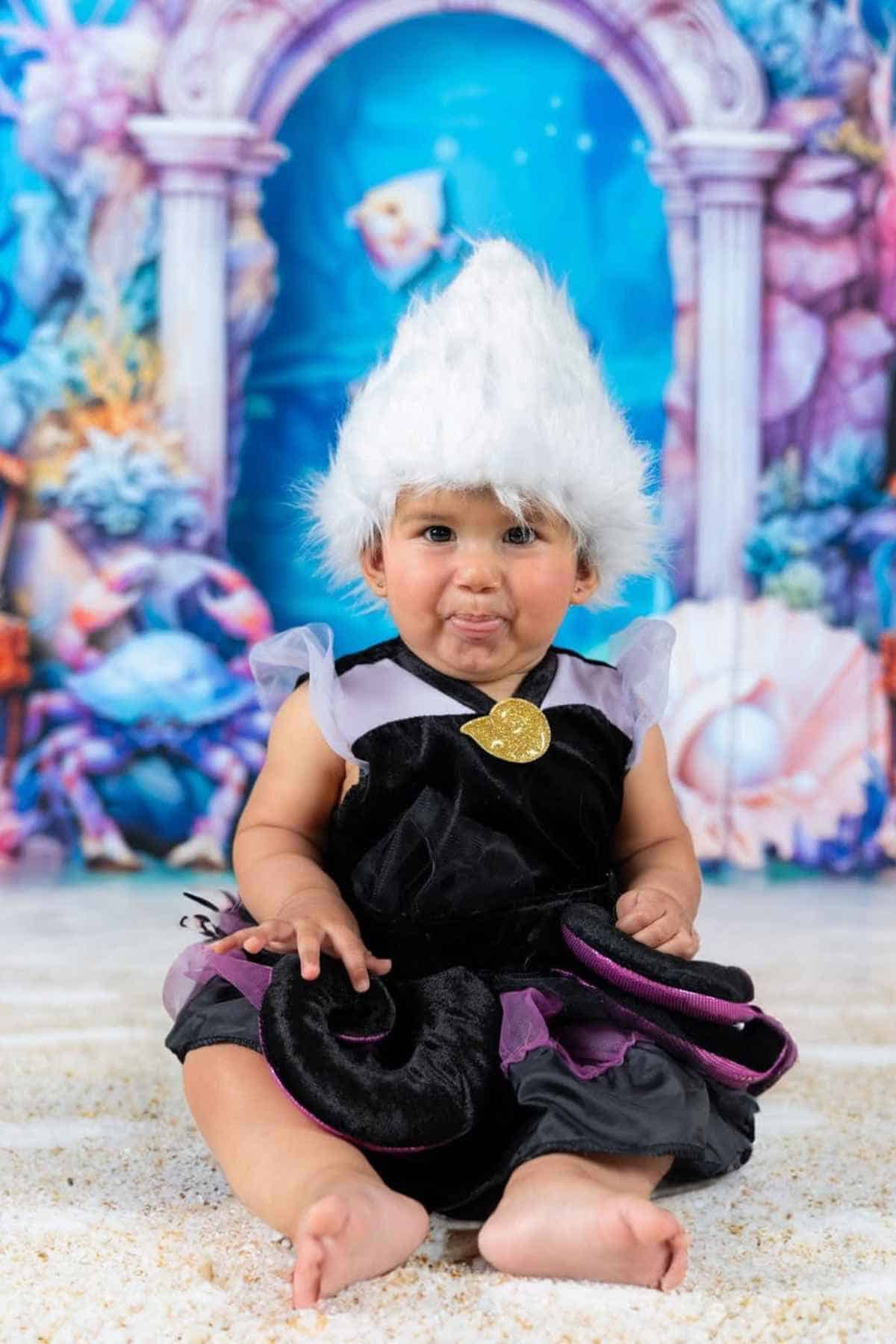 A baby in a black and purple costume with a white wig sits on the floor in front of an underwater-themed backdrop.