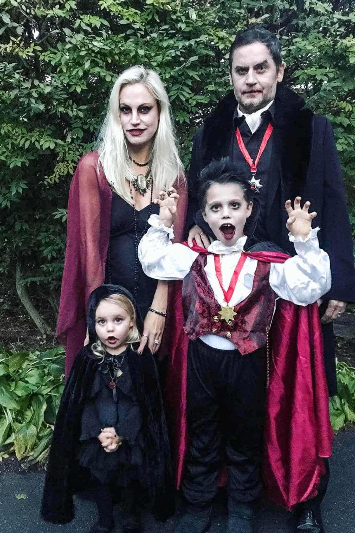 A family of four dressed in vampire costumes stands outdoors, with the adults and children wearing capes, makeup, and fangs.
