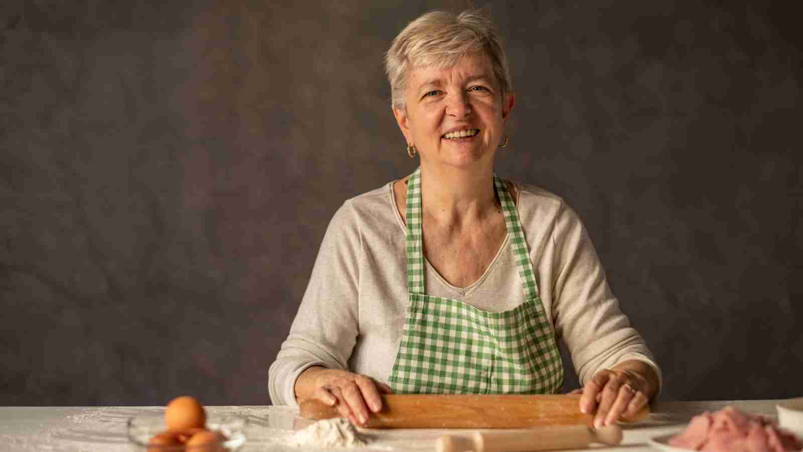A smiling woman with short gray hair, wearing a green and white checkered apron, is at a table with flour, eggs, and a rolling pin.