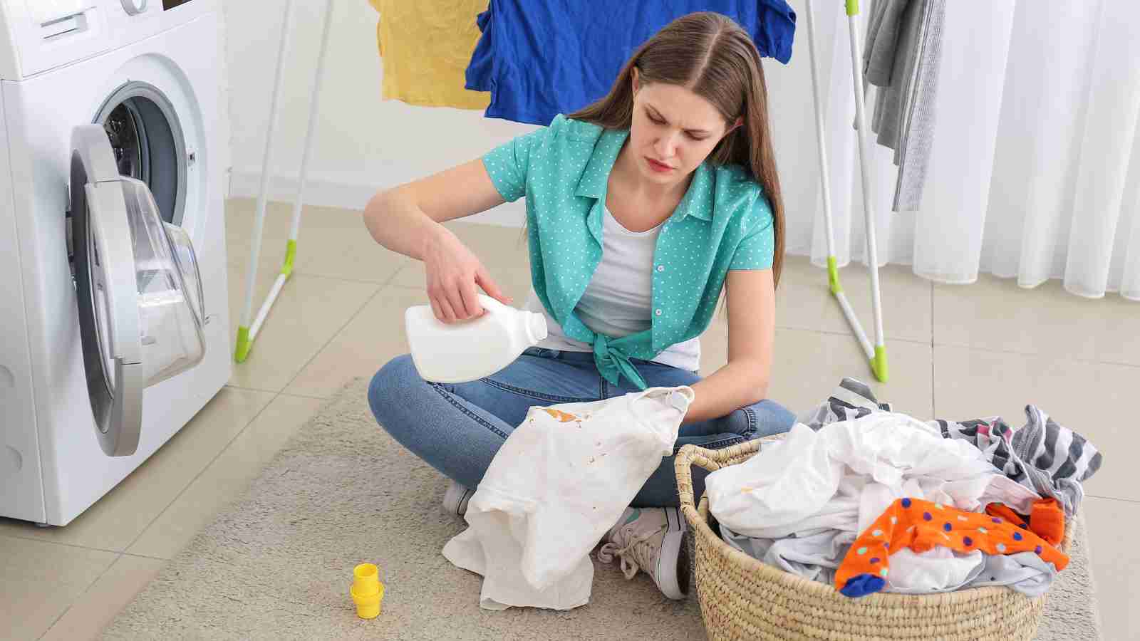 A young woman sits on the floor near a washing machine, treating a stain on a piece of clothing before washing it.
