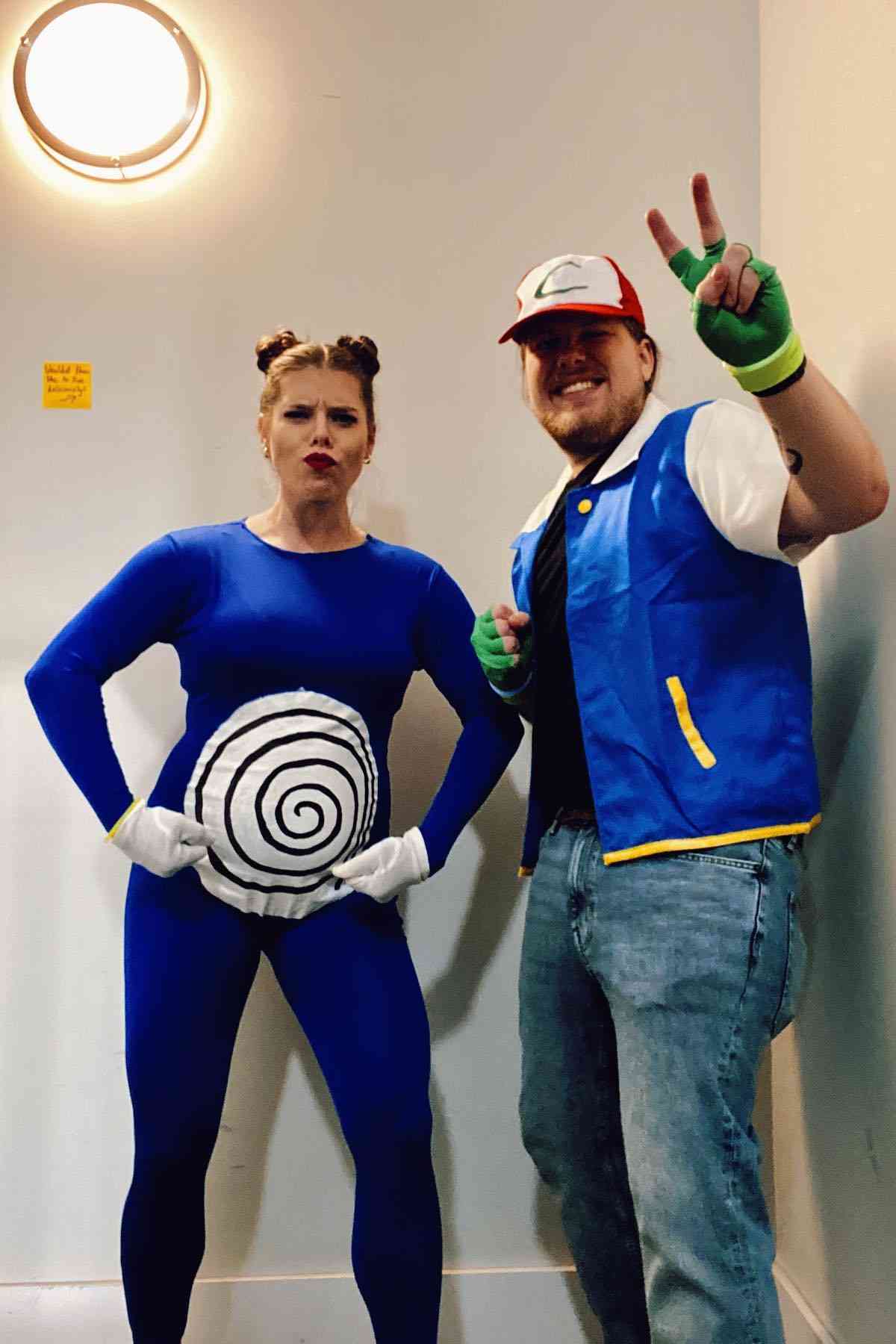 Two adults in costumes: one dressed as the Pokémon Poliwrath with a blue bodysuit and swirl, and the other as Ash Ketchum in a hat, vest, jeans, and gloves, posing indoors.