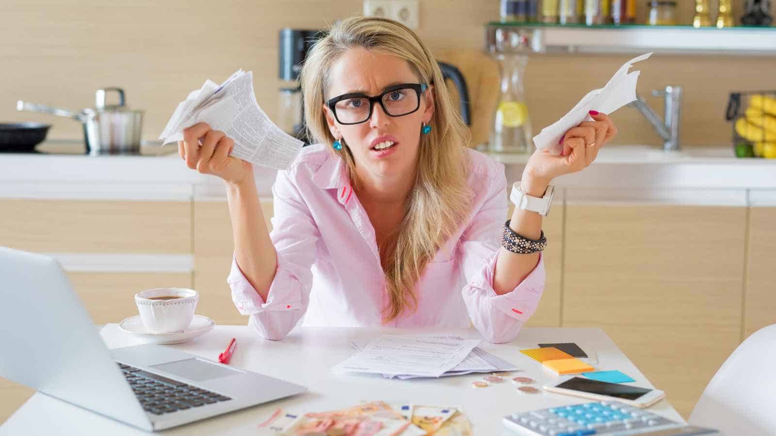A frustrated woman sits at a table, surrounded by bills and money, looking stressed about her finances.