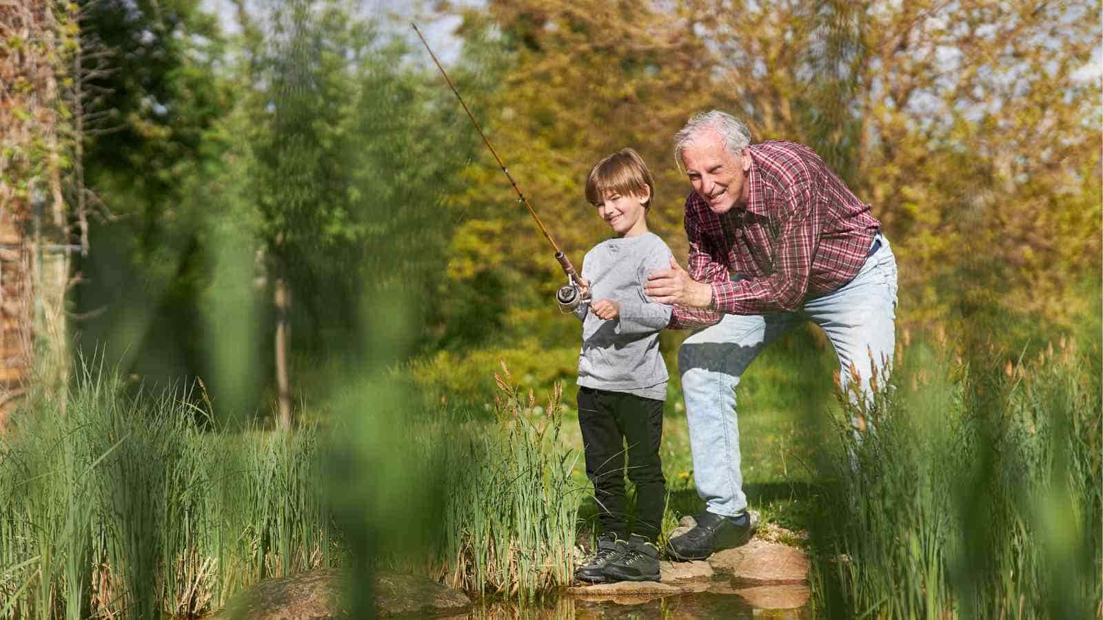 A grandfather assists his grandson as he learns to fish by a pond in a sunny, wooded area.