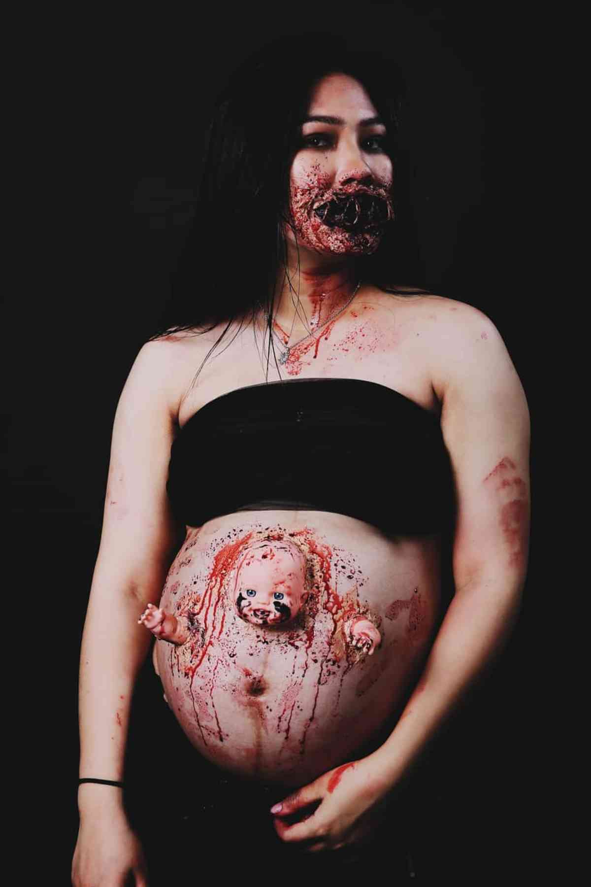 A person with special effects makeup appears bloodied, with a baby doll emerging from their pregnant belly and distorted facial features, against a black background.