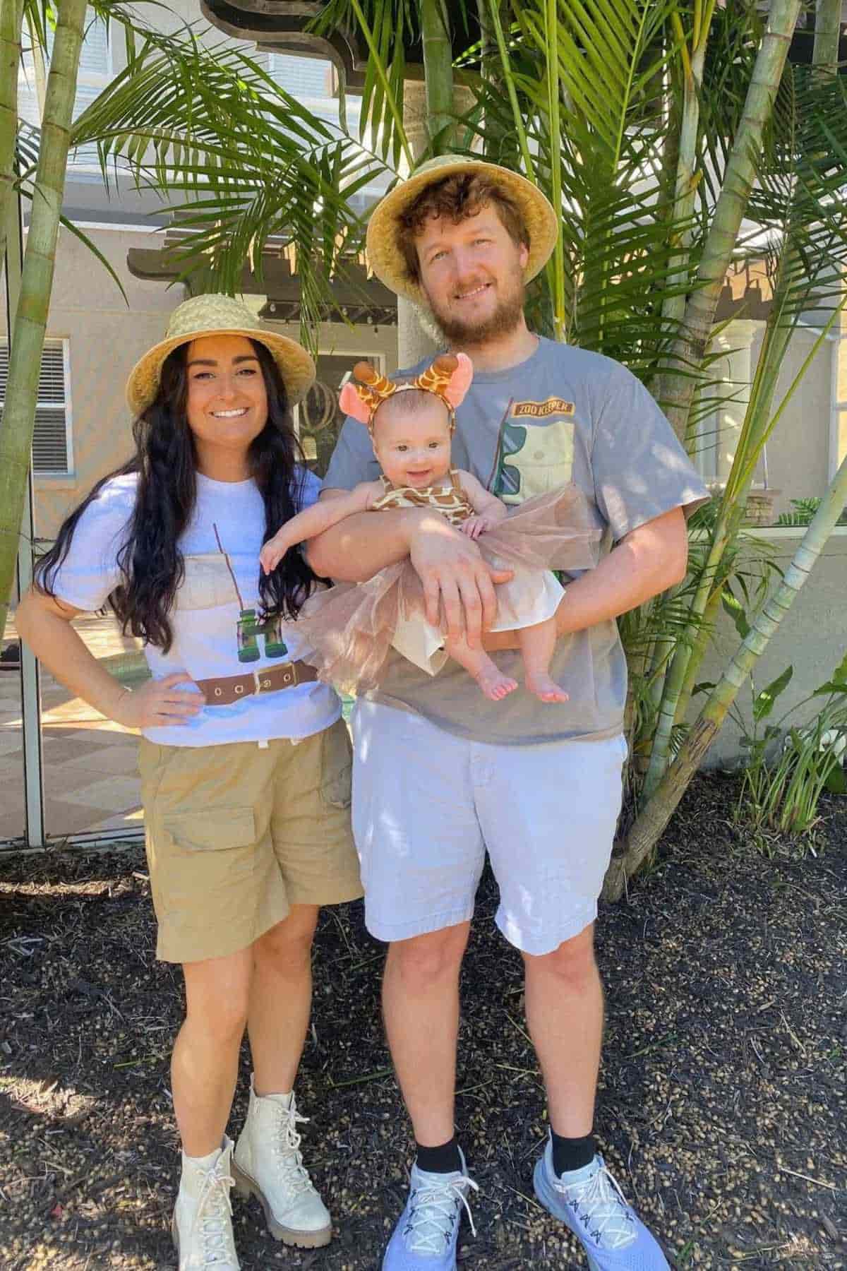 A woman, a man, and a baby dressed in safari-themed and animal-themed outfits stand together outdoors in front of tropical plants.