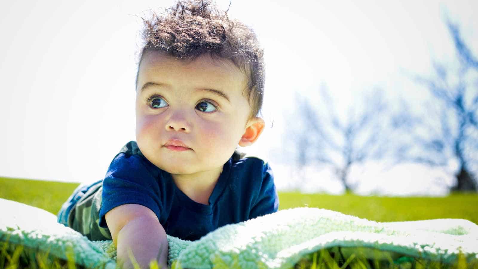 A baby lying on his back on a green mat outside, in broad daylight.