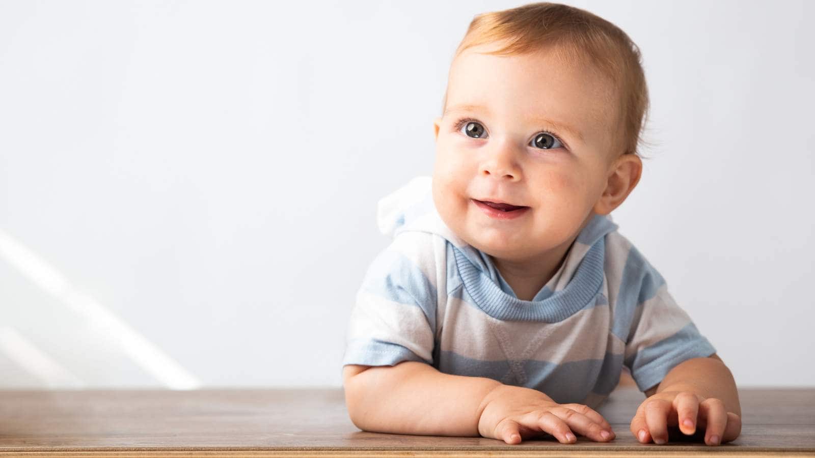 A baby boy wearing a striped blue shirt is crawling on a wooden surface.