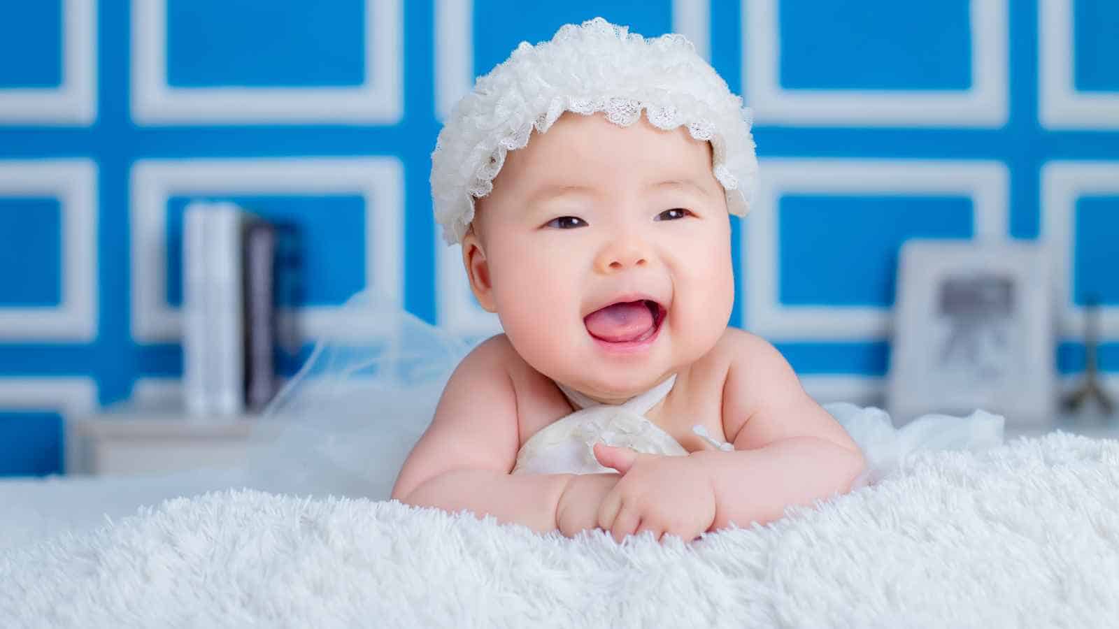 A baby wearing a white headband lies on her back on a white sheet, with a blue patterned background.