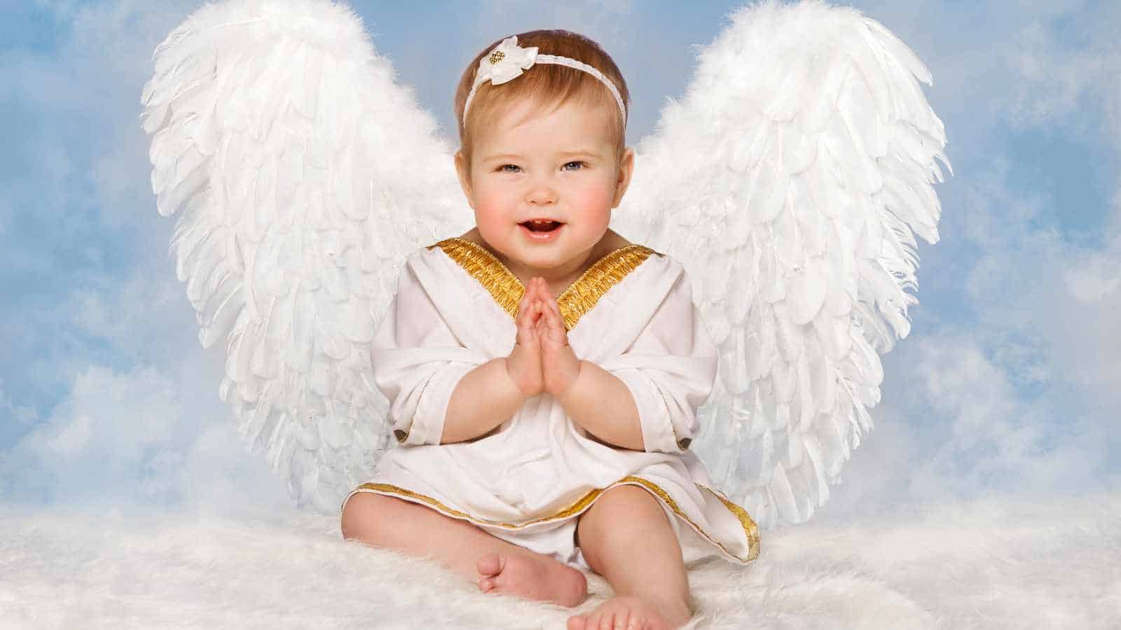 A baby wearing an angel attire from clothe, wings and white headband, posing with a prayer hand behind in a cloudy background.