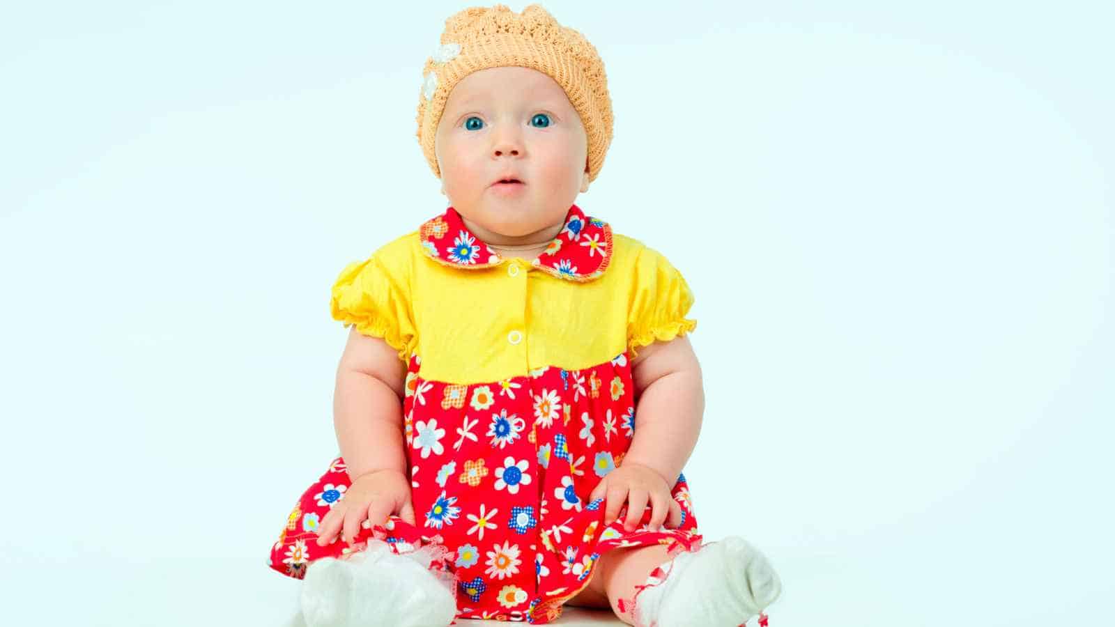 A baby sitting in front of a white background, wearing a red and yellow floral dress and a crocheted hat.