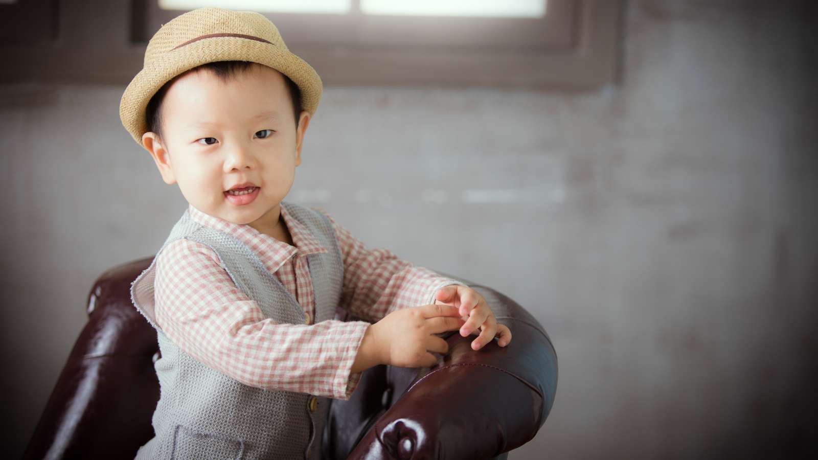 Young child wearing a straw hat and plaid shirt stands next to a brown leather chair, smiling indoors in soft natural light.