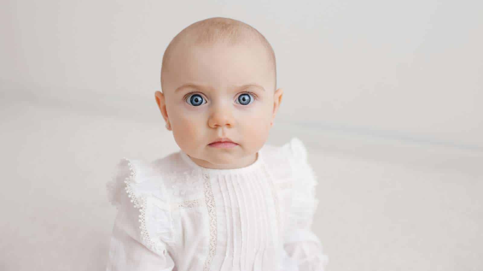 A baby with blue eyes and light hair wearing a white outfit sits against a neutral background, looking directly at the camera.