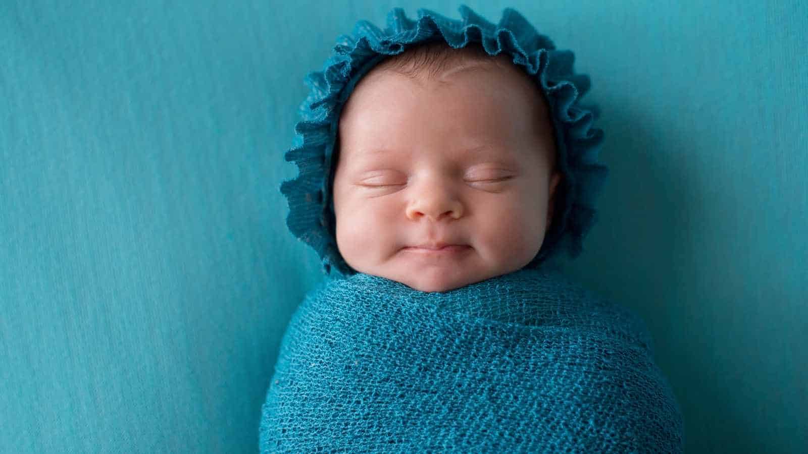 A newborn baby wrapped in a blue blanket with a matching bonnet, lying on a blue background, eyes closed and a slight smile on their face.