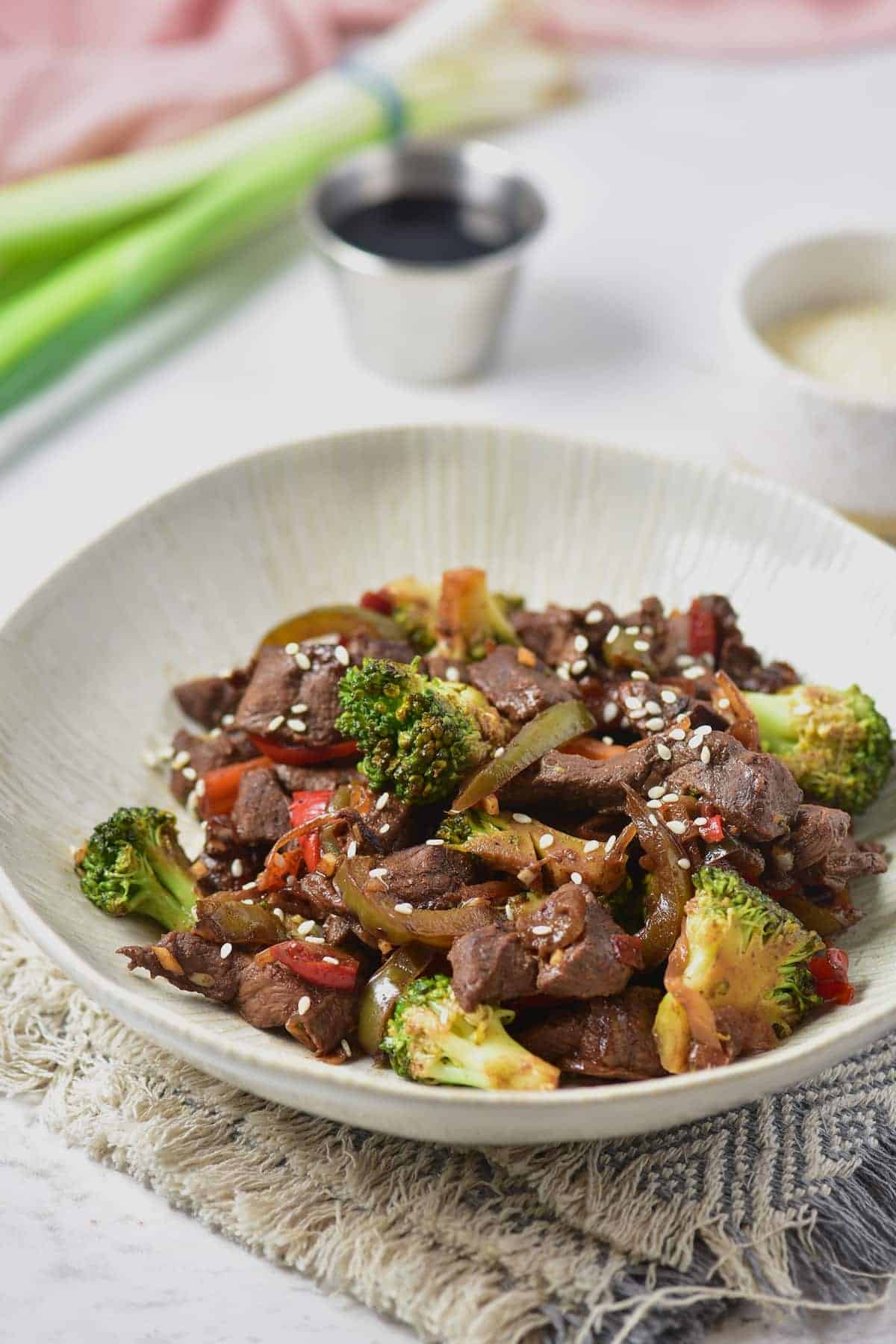 Ready-to-serve venison stir fry plated on a white textured dish, with a steel cup and a bundle of leeks nearby.