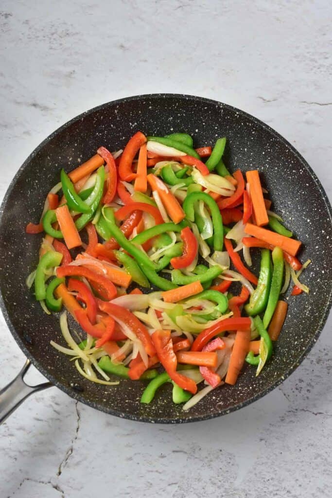 Ingredients like sliced carrots, sweet peppers, and onions sautéed in a pan.