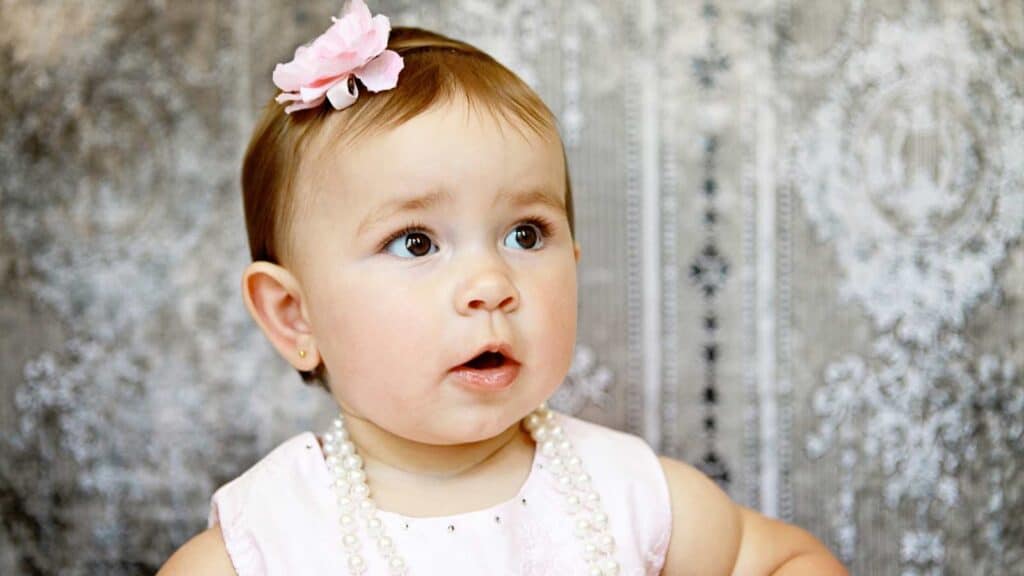 A baby girl wearing fancy necklace, earing and a pink bow looking sideways.