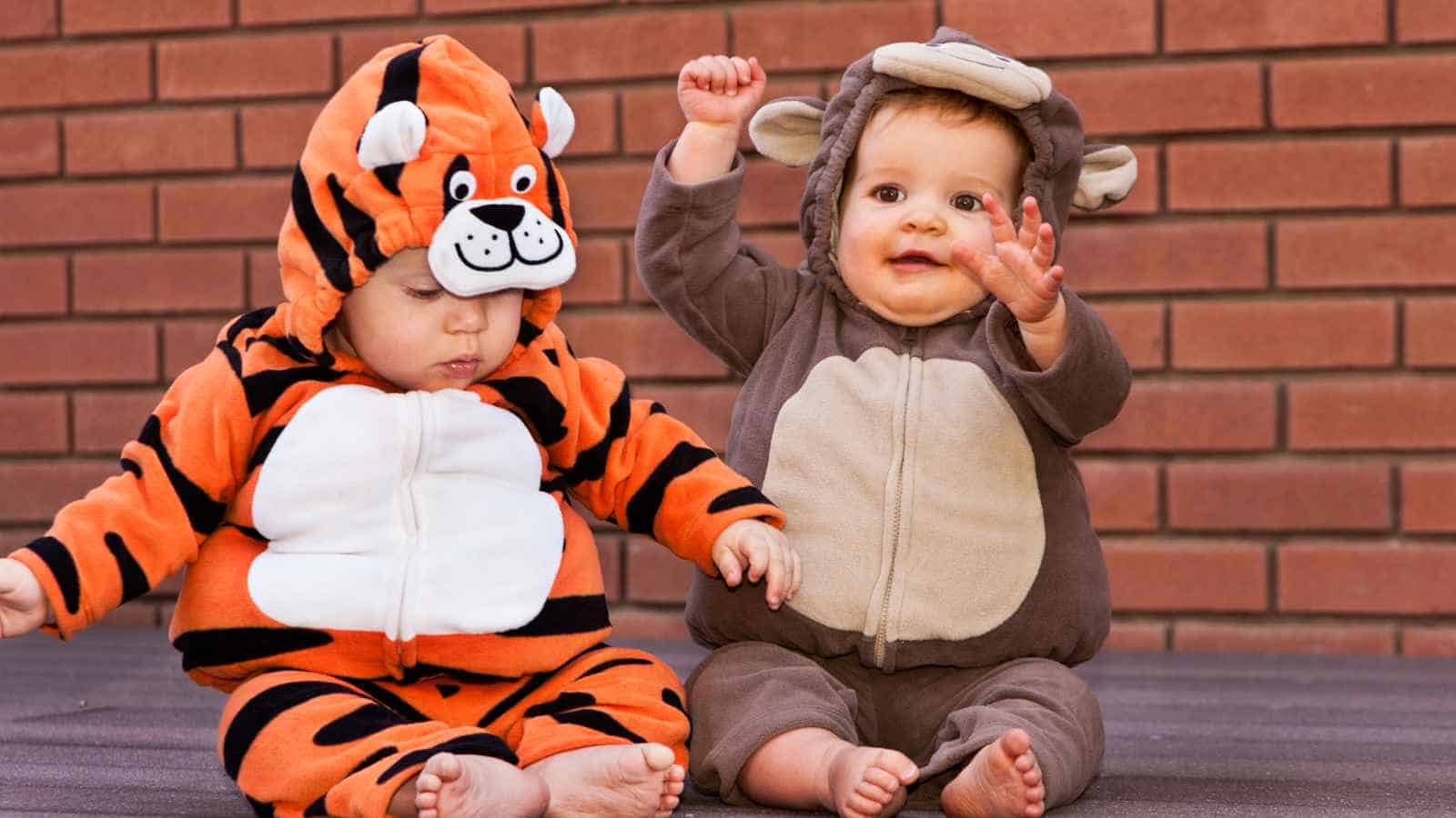 Two babies sit side by side in animal costumes, one dressed as a tiger and the other as a monkey, against a brick wall background.
