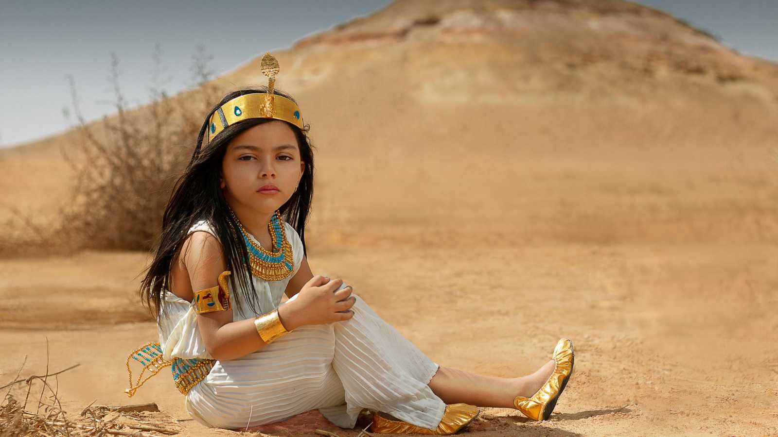 A young girl in ancient Egyptian-style costume sits on sandy ground in front of a barren hill, wearing gold accessories and a beaded necklace.
