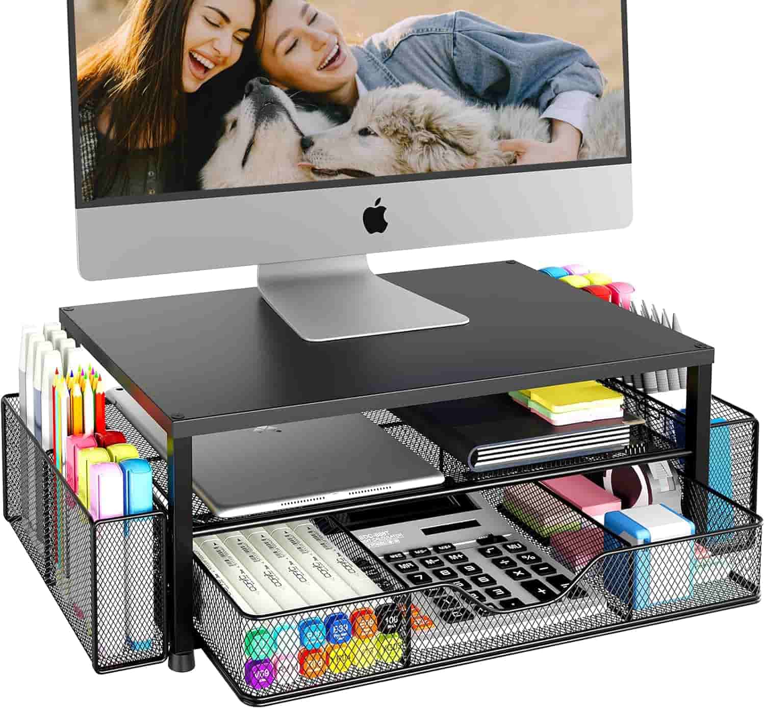 A computer monitor on a black mesh desk organizer holding office supplies like pens, sticky notes, a calculator, and notebooks, with two people and two dogs shown on the screen.