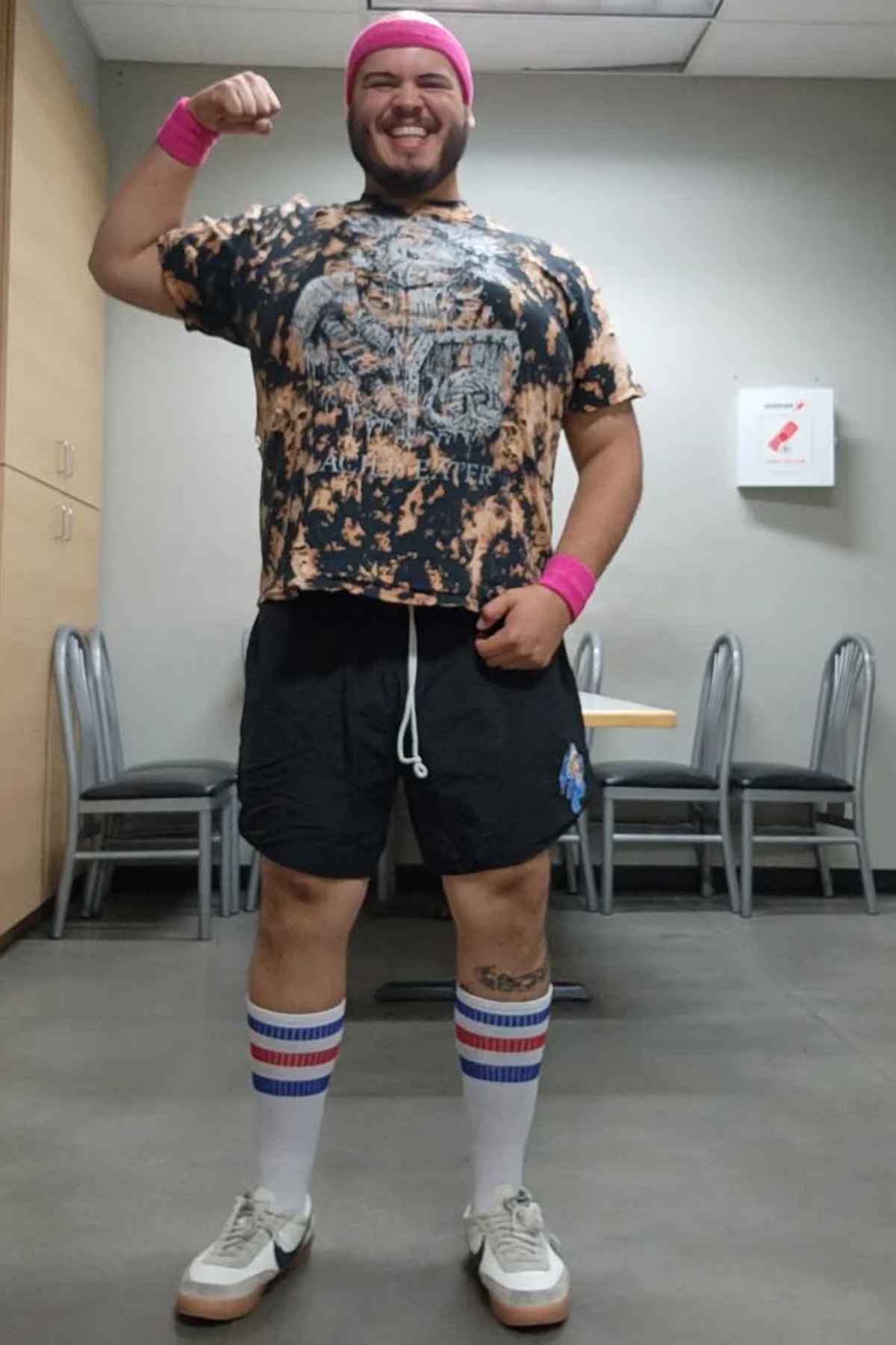 A person is standing indoors, flexing one arm and smiling. They are wearing a pink headband and wristbands, a tie-dye shirt, black shorts, tall striped socks, and white shoes.