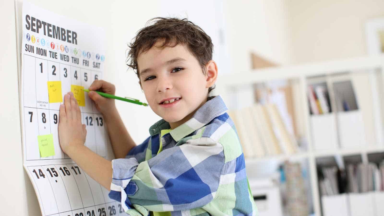 Young boy writing on a large wall calendar for September with a green pencil, standing indoors and looking at the camera.