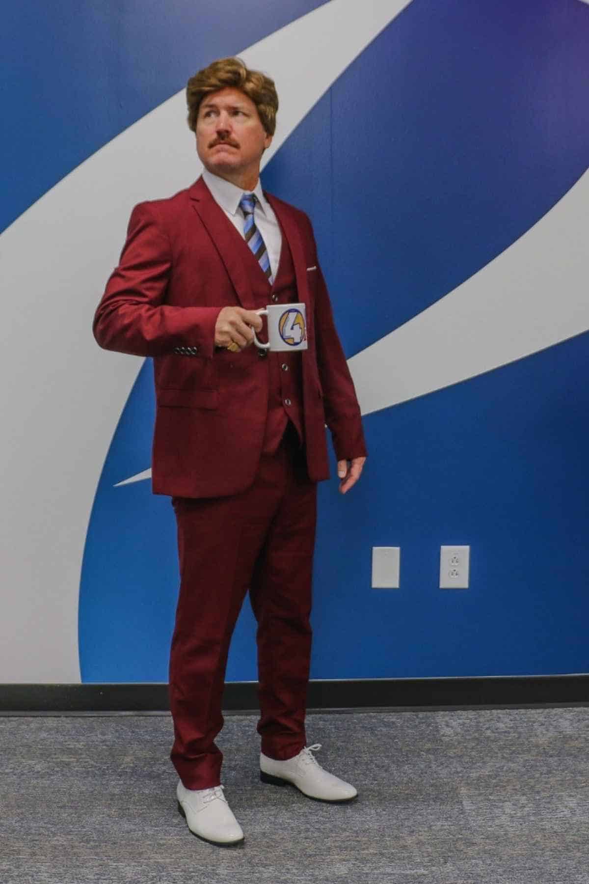 A man in a maroon suit holding a coffee mug stands in front of a blue and white wall, looking to the side.