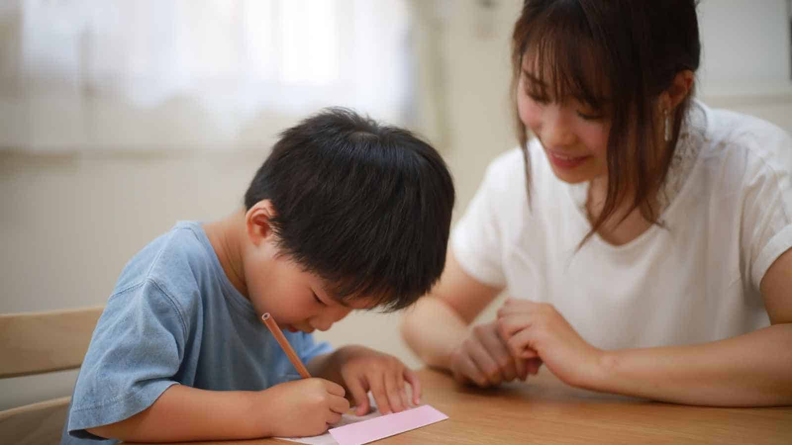 A young boy writes on a piece of paper at a table while an adult woman sits beside him, watching and smiling.