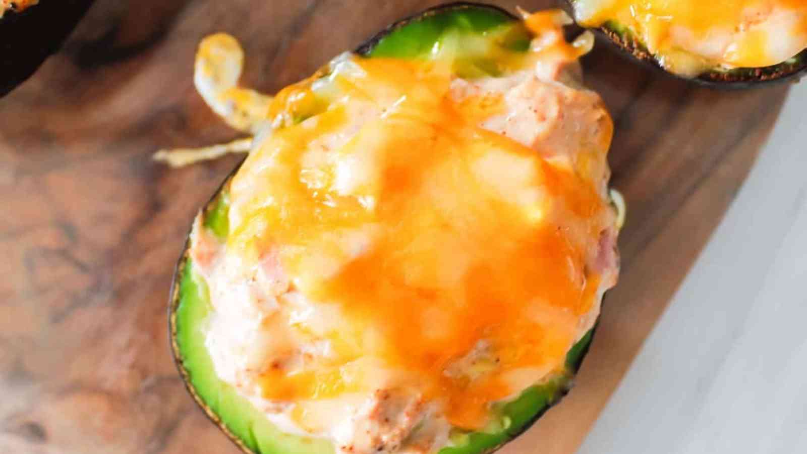 Halved avocado filled with a creamy mixture and topped with melted cheddar cheese, placed on a wooden surface.